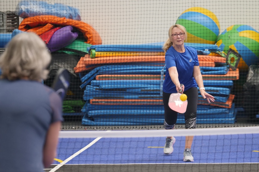 A woman in a blue shirt, hitting a yellow ball over a net with a paddle. She is playing pickleball against an opponent. 