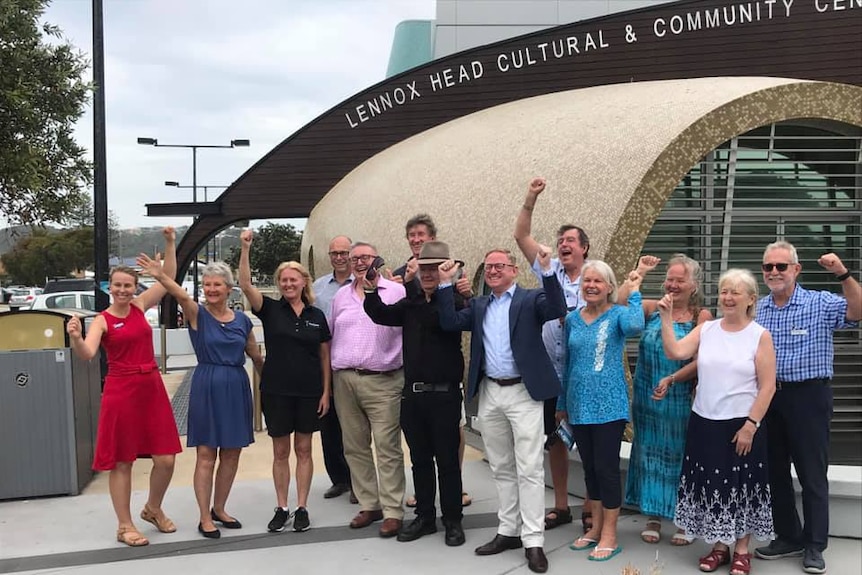 A group of people raise their hands and cheer in front of the Lennox Head Community Centre.