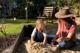 A woman in a hat sits with a toddler in a sandpit. 