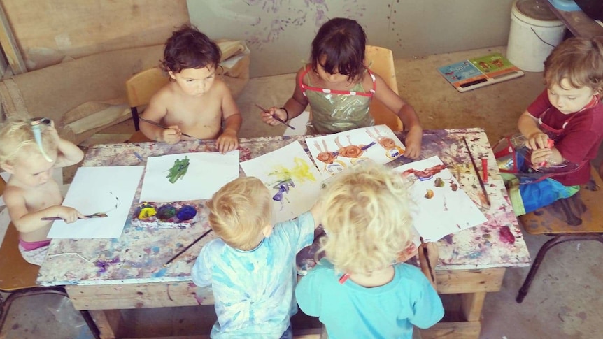 Six young children sitting around a table painting in a messy paint splattered home class room