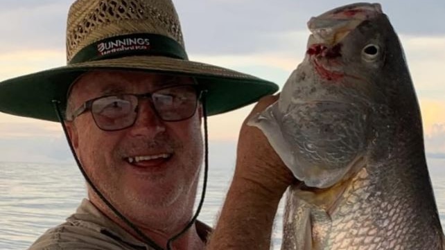 A man wearing a wide-brimmed hat and glasses, smiles for a photo as he holds aloft a fish.