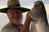 A man wearing a wide-brimmed hat and glasses, smiles for a photo as he holds aloft a fish.