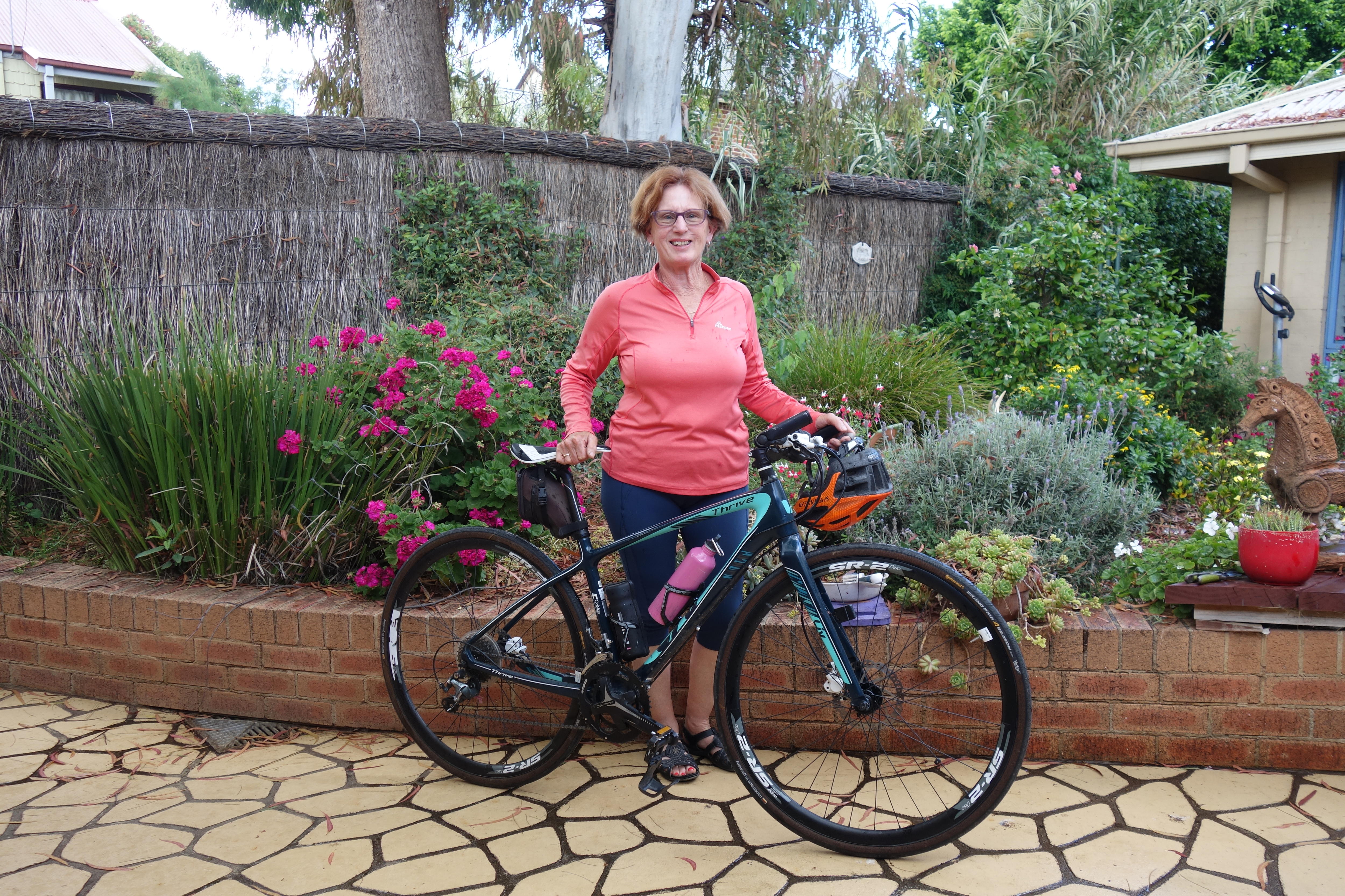 A woman stands behind her bike in front of a flower garden.