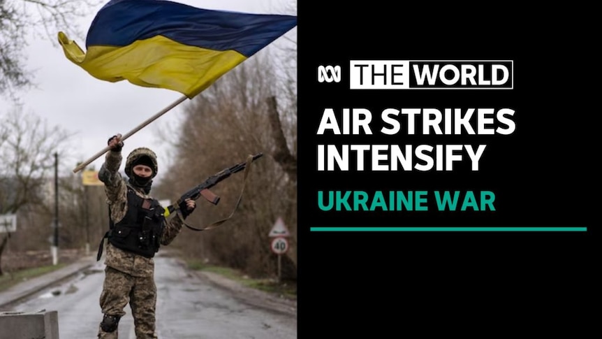 Air Strikes Intensify, Ukraine War: A man wearing military fatigues waves a Ukrainian flag while holding an automatic rifle.