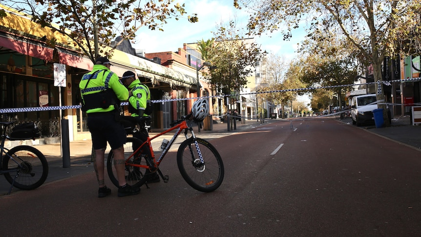 Two police officers with bicycles stand on a street with police tape