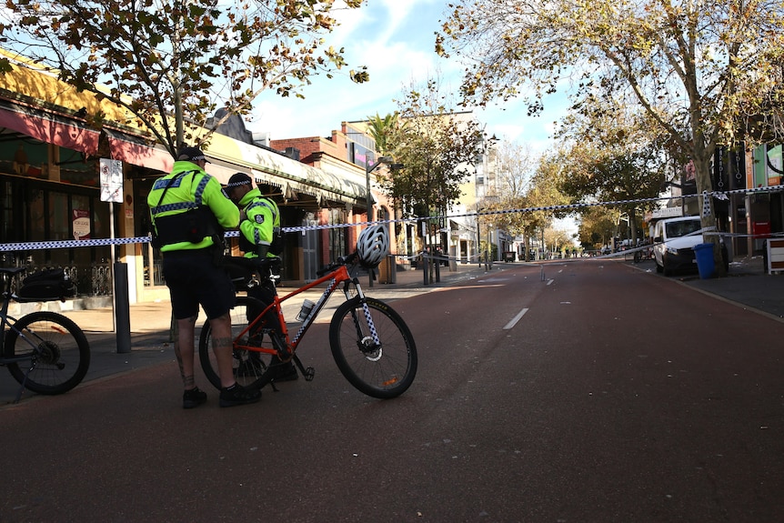 Two police officers with bicycles stand on a street with police tape