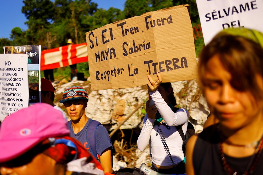 Protestors holding signs written in Spanish stand in sunlight. 