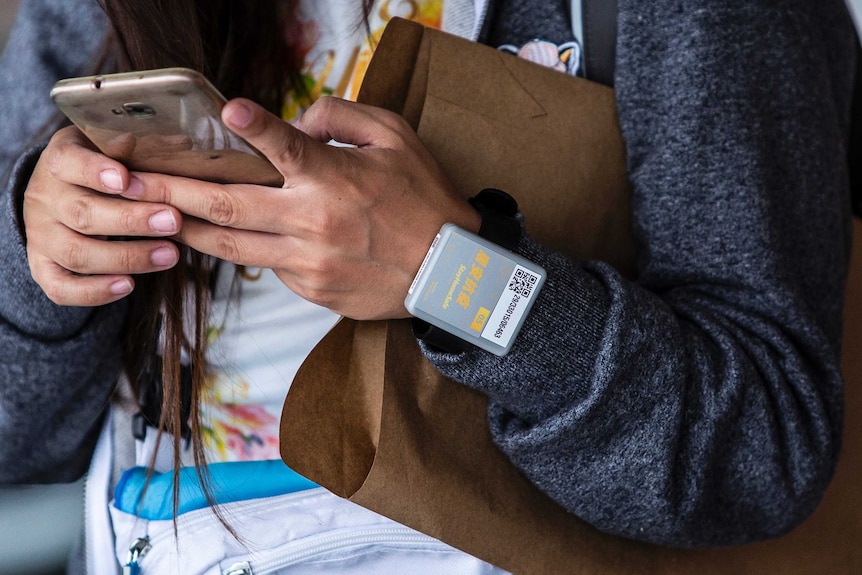 A woman holds a mobile phone, on her wrist is a grey square plastic device on a wristband.