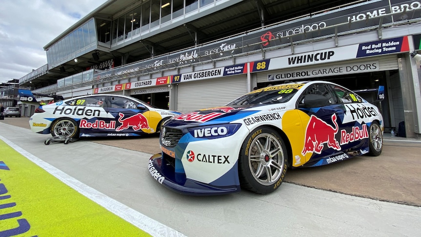 Two Red Bull Holden Commodore Supercars are parked in front of a pit garage