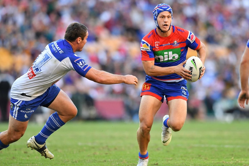 Kalyn Ponga, with his headgear on, looks upfield while carrying the ball. A defender is approaching him.