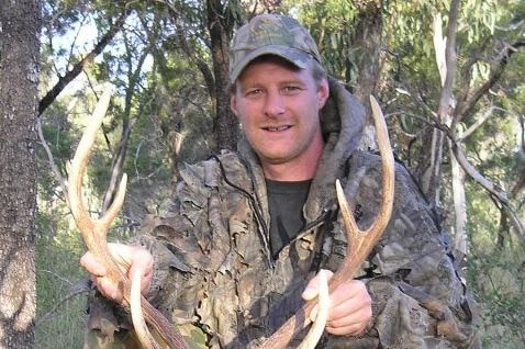 Bryce Airs in hunting clothing poses in the bush with the body of a deer.