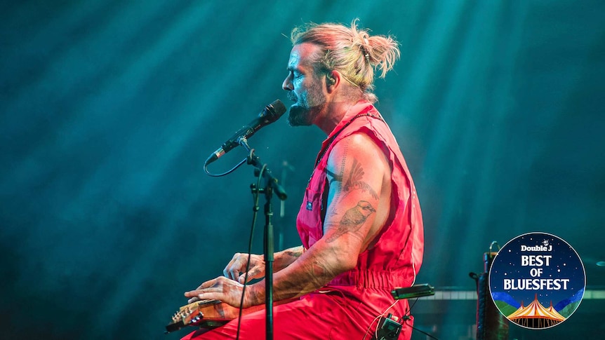 Side view photograph of Xavier Rudd on stage. He is wearing a red onesie, with blonde hair, singing and playing slide guitar