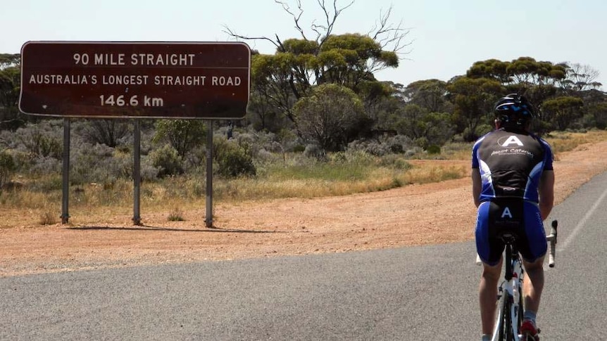 Dave cycles by a sign saying Australia's longest straight road