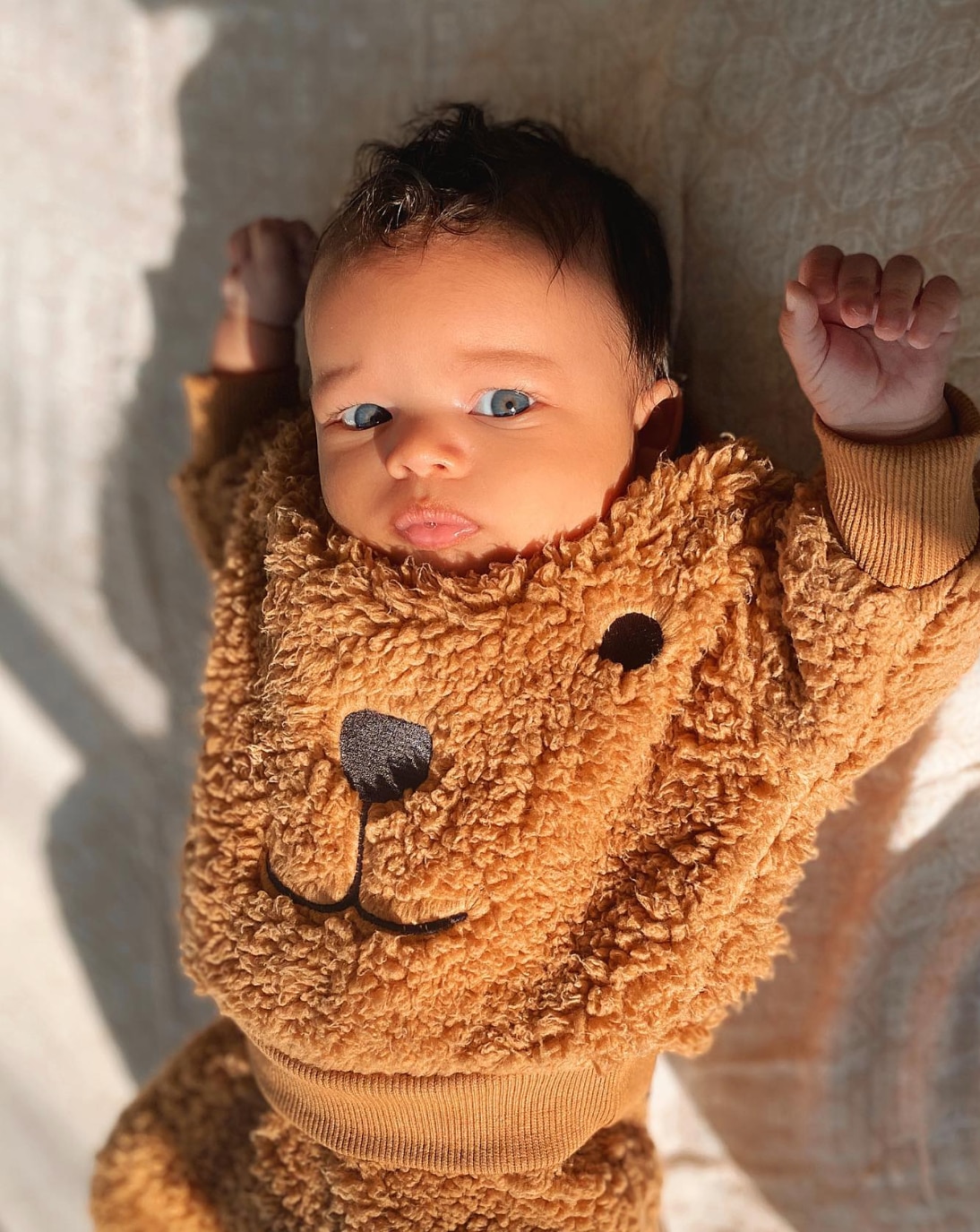 A baby boy in a brown teddy outfit is lying down stretching out.