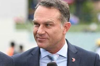 Michael Slater – dressed in a suit and with short, greying hair – stands in an outdoor sports venue.