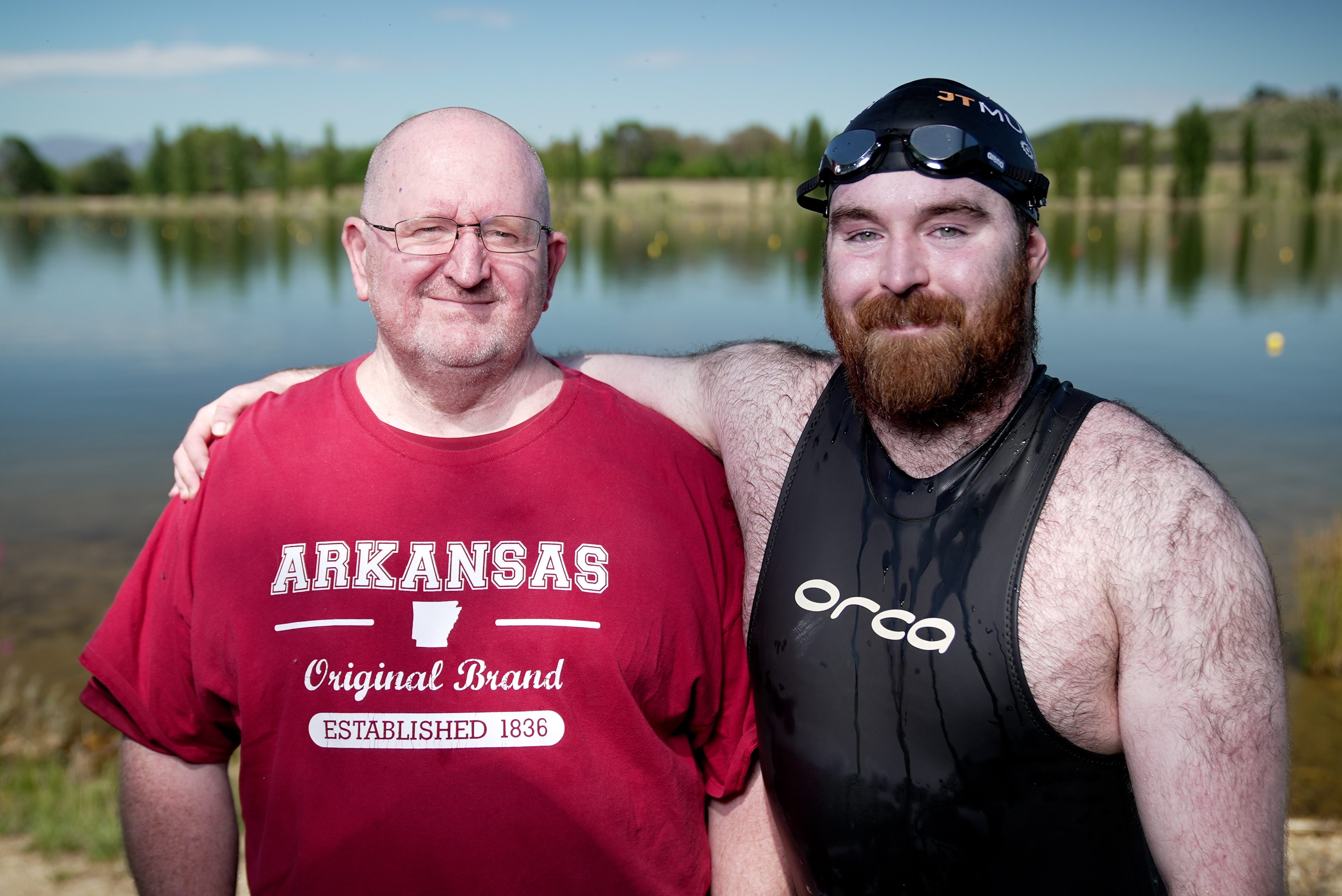 a man in a wetsuit next to an older man in a red top on the shores of lake burley griffin