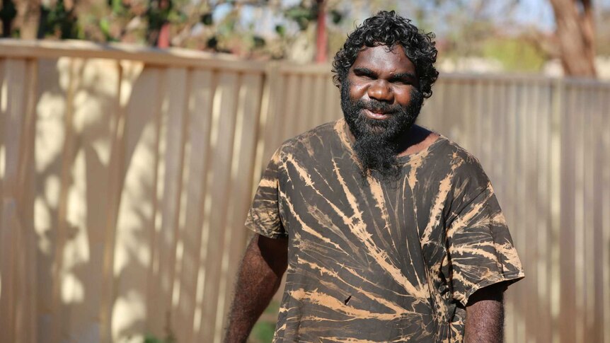 An Aboriginal man with a black and brown patterned shirt, black beard, and curly black hair, stands smiling.