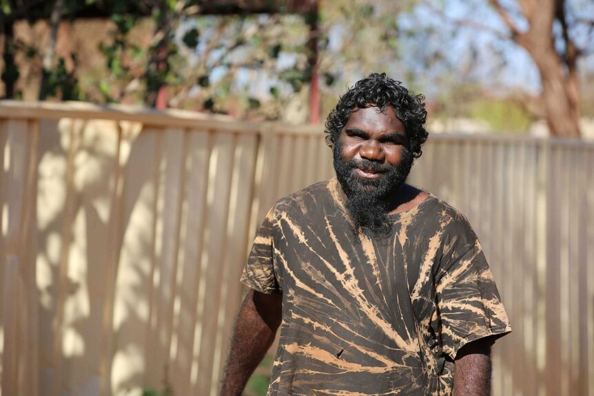 An Aboriginal man with a black and brown patterned shirt, black beard, and curly black hair, stands smiling.
