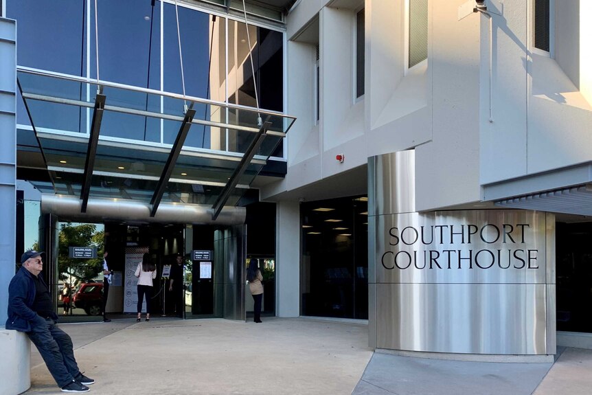 A man waits at the entry to a modern, glass and steel building with Southport Courthouse written on a shiny round pillar.