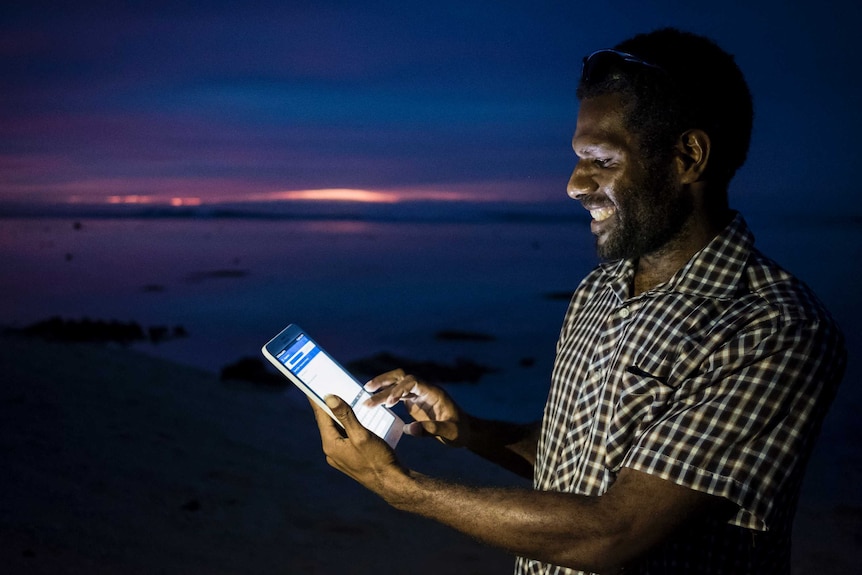 A dark-skinned man wearing a checked shirt holds a tablet as the sun sets.