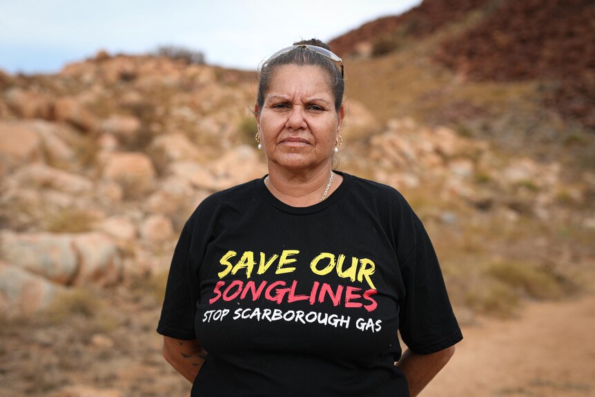 Raelene Cooper wears a shirt that reads 'save our songlines' and is standing outside in front of rock formations