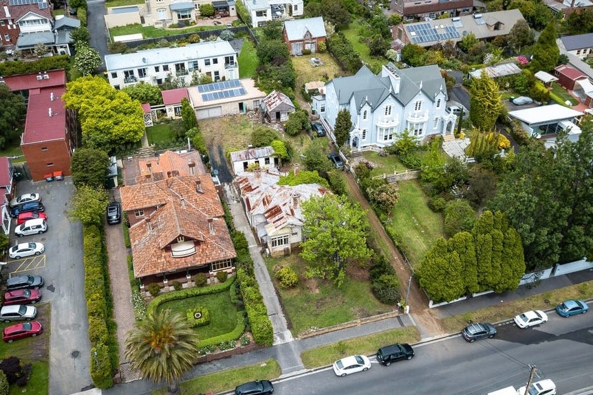 An aerial shot of a dilapidated house next to mansions in a leafy suburb