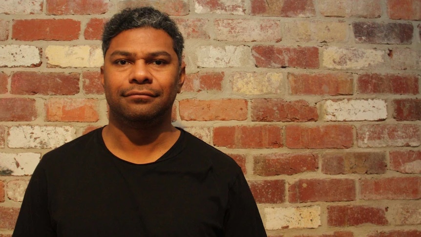 A man with black hair wearing a black shirt standing in front of a brown brick wall.