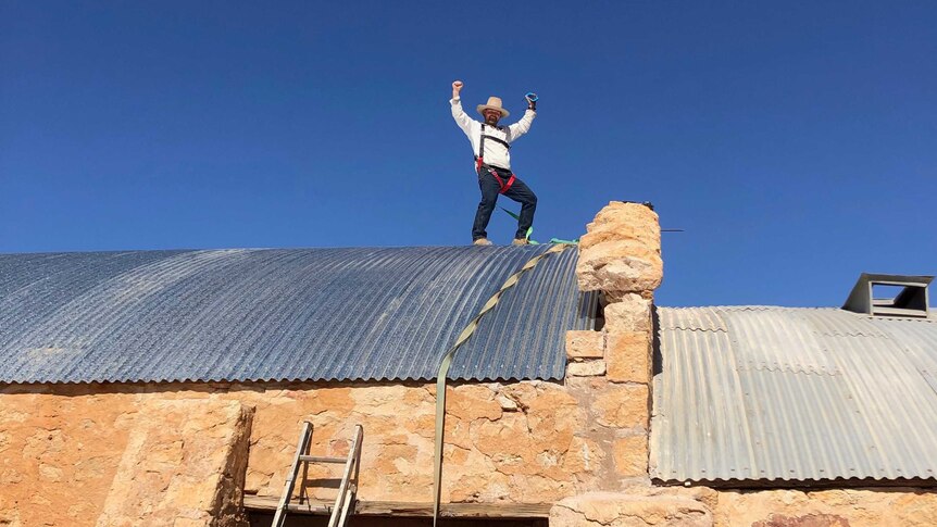A man in a white shirt and large hat celebrates on top of a historic woolshed.