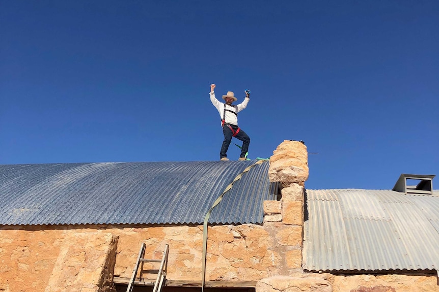 A man in a white shirt and large hat celebrates on top of a historic woolshed.