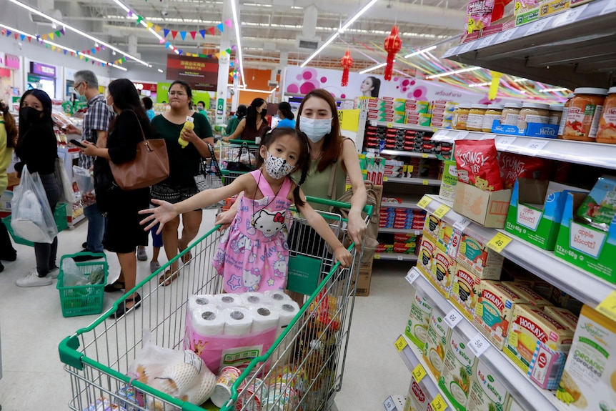 A child stands in a shopping trolley in a supermarket being pushed by her mother wearing a facemask.