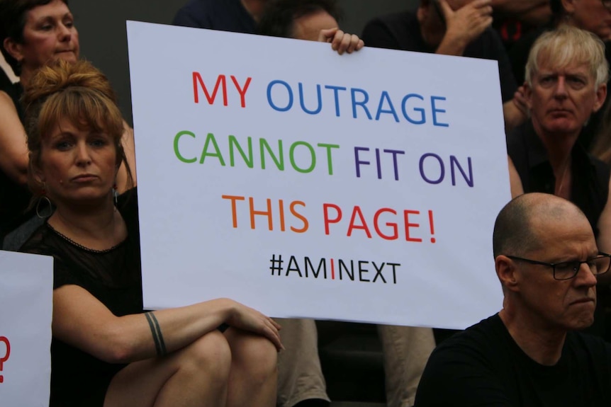 A woman holds a sign reading, "My outrage cannot fit on this page! #AmInext".