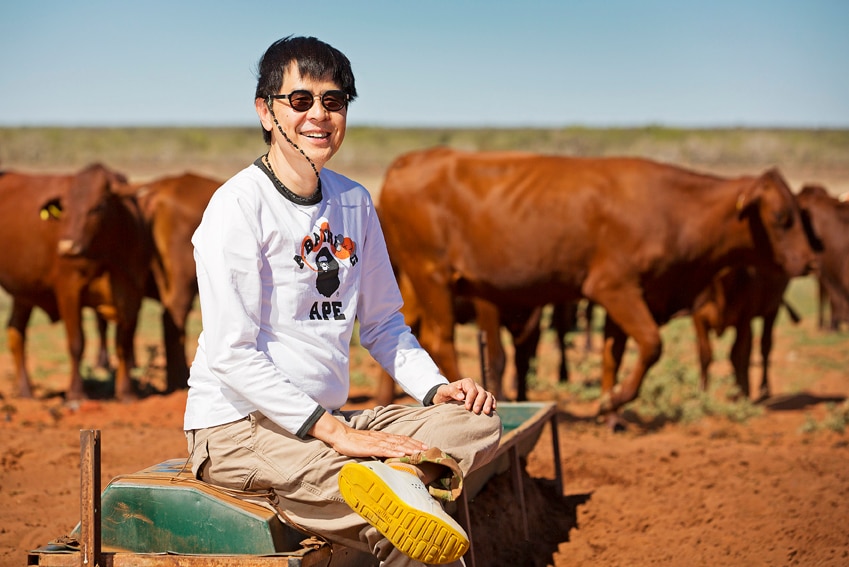 A man sits with cattle and red dirt behind him.