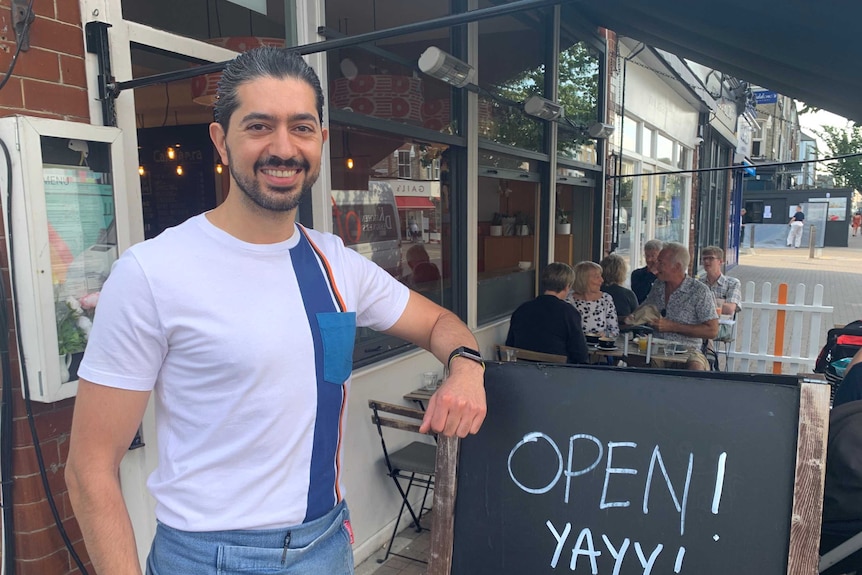 A man with black hair and a beard rests his arm on a sign saying 'Open Yay'