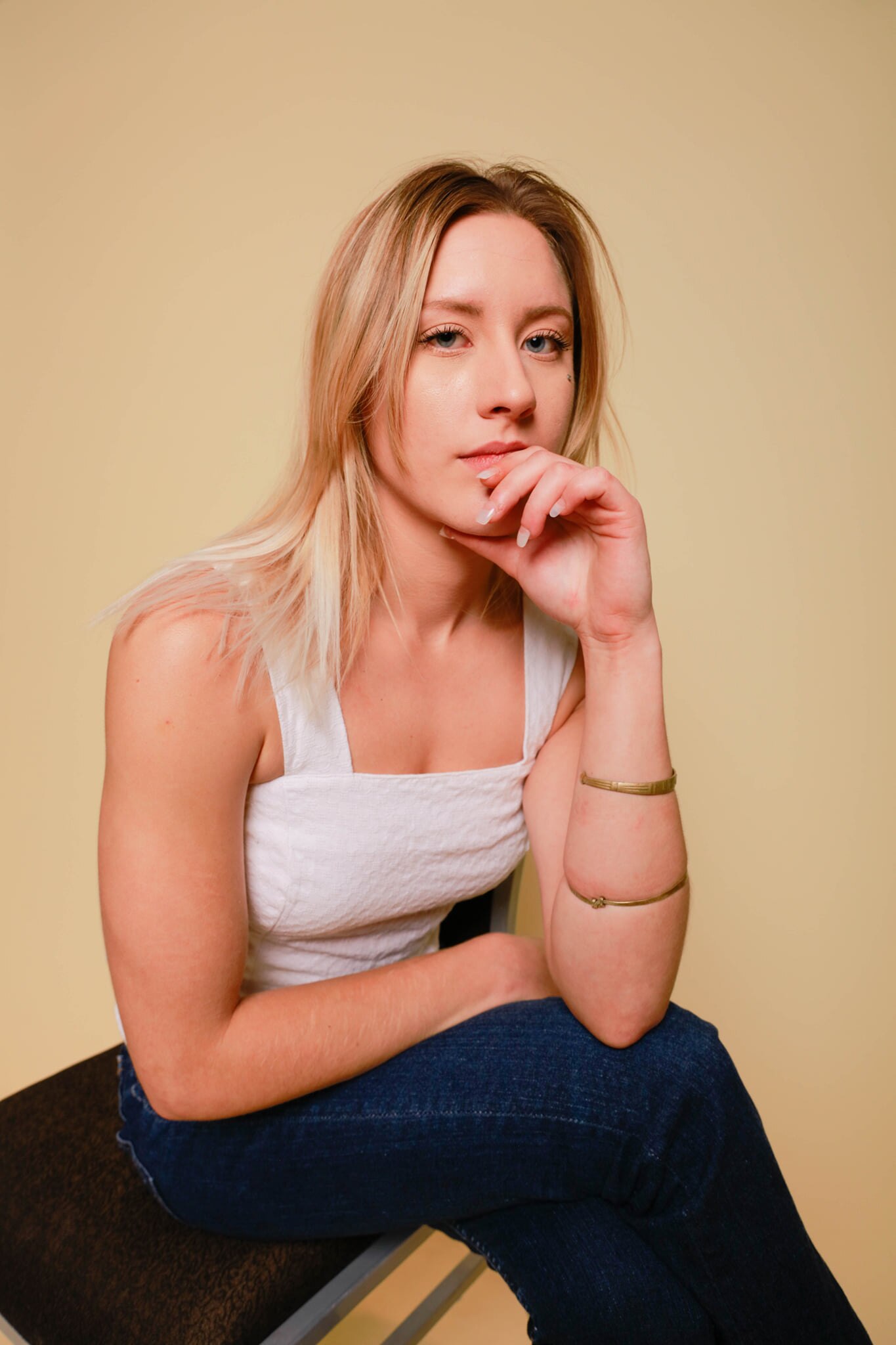 A portrait shot of a young blonde woman sitting down wearing a white top with her head resting on her hand.