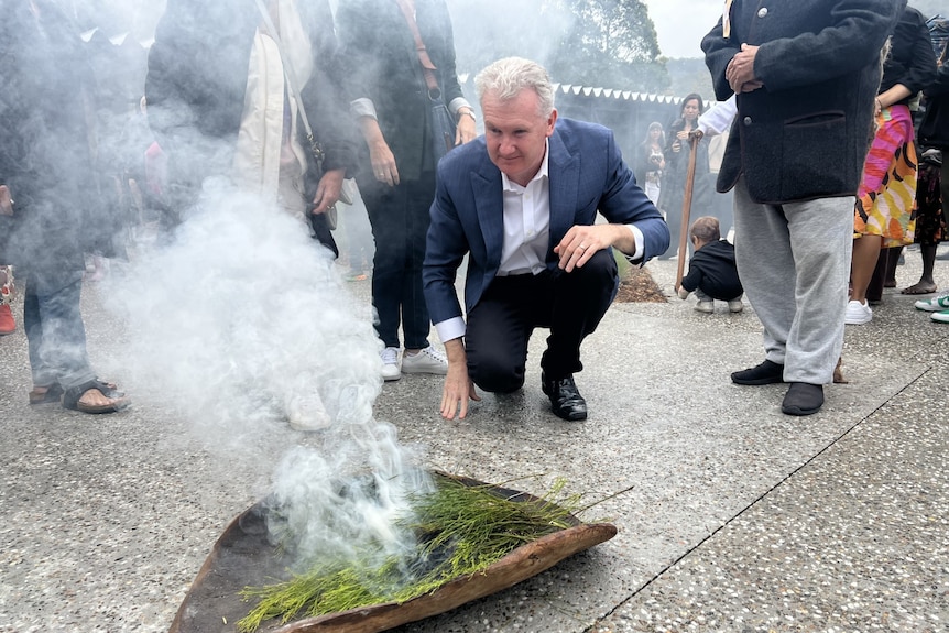 Minister Tony Bourke is bending down in front of a coolaman with leaves and smoke drifting upwards