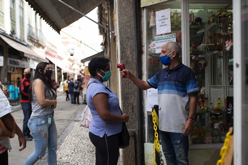 A man points a thermometer gun and a woman's forehead as she stands in line to get into a store. Everyone is in masks.