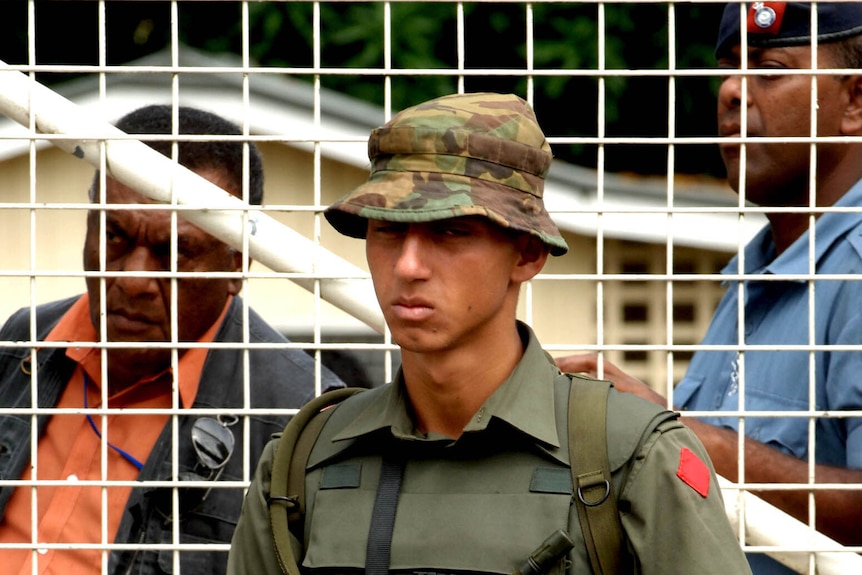 Ratu Meli Bainimarama stands at a fence with a gun. He is dressed in a military uniform
