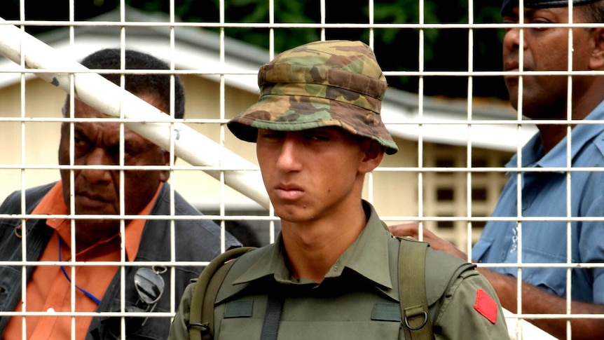 Ratu Meli Bainimarama stands at a fence with a gun. He is dressed in a military uniform