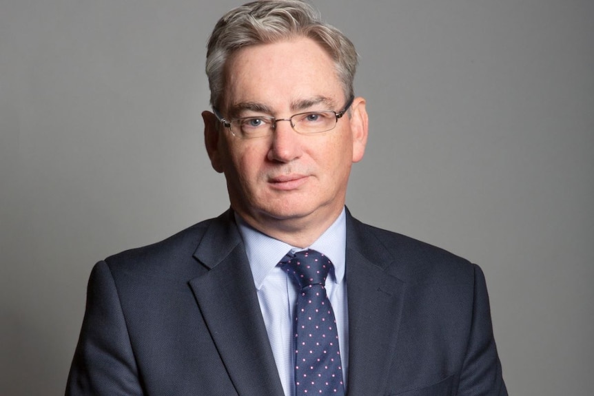 A man with grey hair wears a suit and glasses.