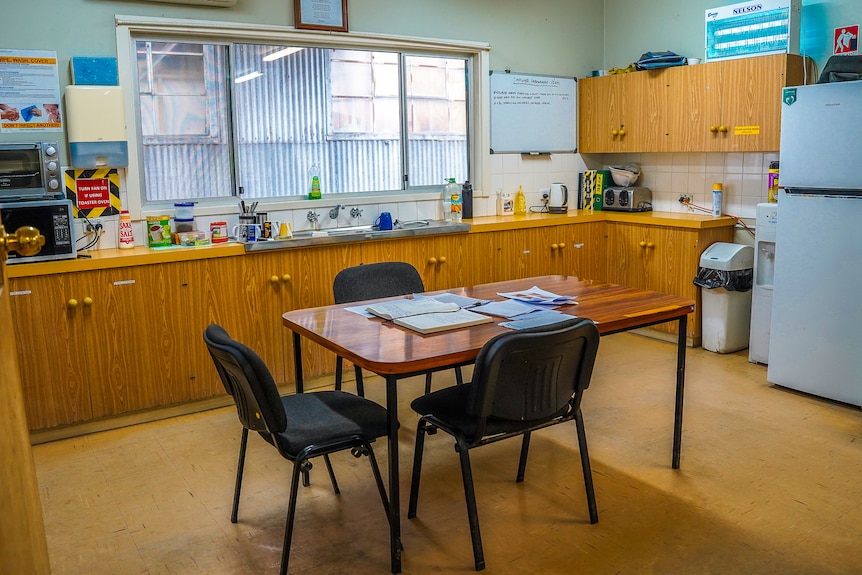 A bright, well stocked lunch room with a central table surrounded by chairs, a fridge, benches and a sink  