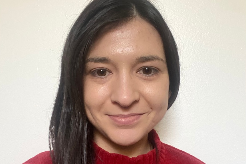 Heidi has long dark hair, is wearing a red top and is standing in front of an empty wall.