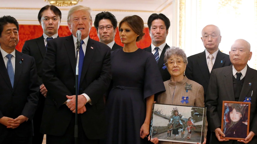 A man stands at a microphone surrounded by Japanese people clutching picture frames