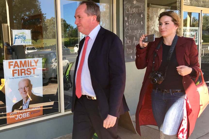 Barnaby Joyce walks in front of Vikki Campion walk past a Family First campaign poster in a shop window