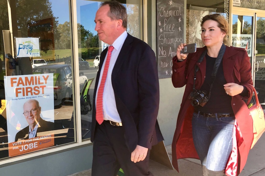 Barnaby Joyce walks in front of Vikki Campion walk past a Family First campaign poster in a shop window.