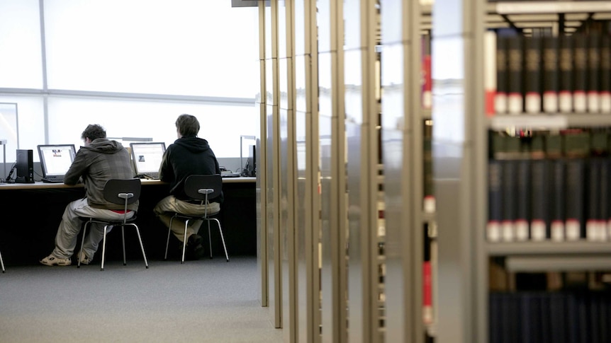 Students work on computers next to shelf stacks in a university library.