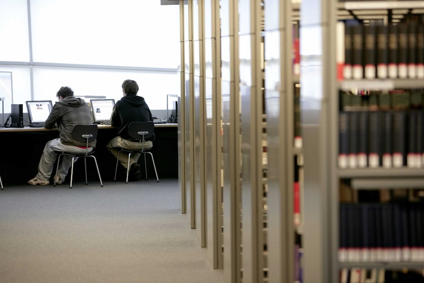 Students work on computers next to shelf stacks in a university library.