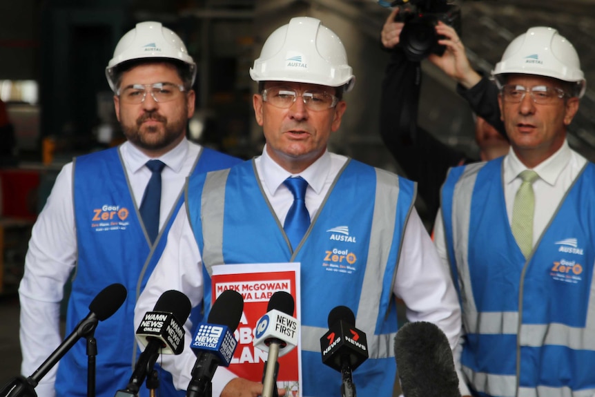 WA Premier Mark McGowan speaking at a media conference wearing a blue hi-vis vest and white hard hat, flanked by two other men.