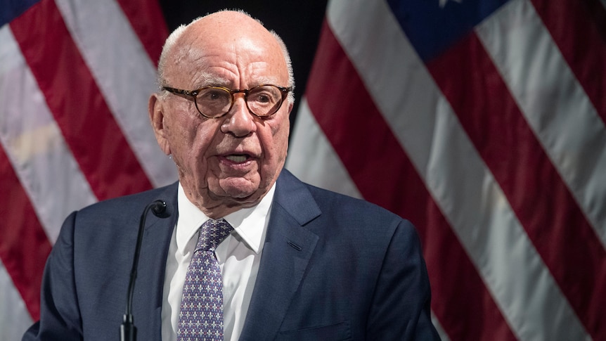 An elderly white man in a suit with horn-rimmed glasses speaks in front of two American flags.