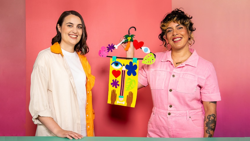 Two smiling women in front of a pink background. One woman holds up a craft artwork she created with a coathanger.