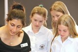 A female school teacher stands with three female Year 7 students looking down at something.